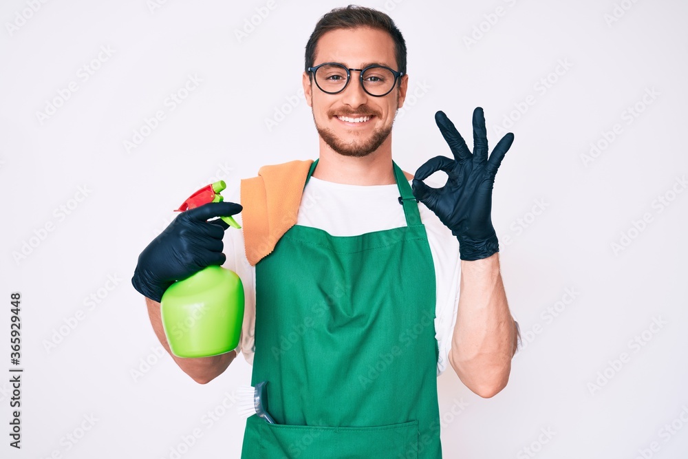 Young handsome man wearing apron holding sprayer doing ok sign with fingers, smiling friendly gesturing excellent symbol Young handsome man wearing apron holding sprayer doing ok sign with fingers, smiling friendly gesturing excellent symbol