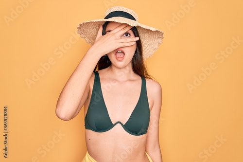 Young beautiful girl wearing swimwear bikini and summer sun hat over yellow background peeking in shock covering face and eyes with hand, looking through fingers with embarrassed expression.
