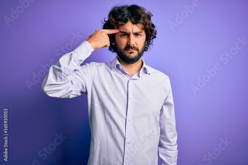 Young handsome business man with beard wearing shirt standing over purple background pointing unhappy to pimple on forehead, ugly infection of blackhead. Acne and skin problem