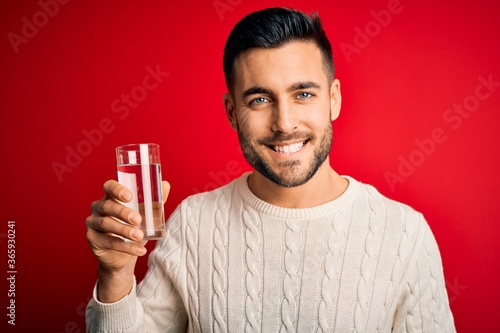 Young handsome man smiling happy and confident. Standing with smile on face holding glass of healthy water to refreshment over isolated red background