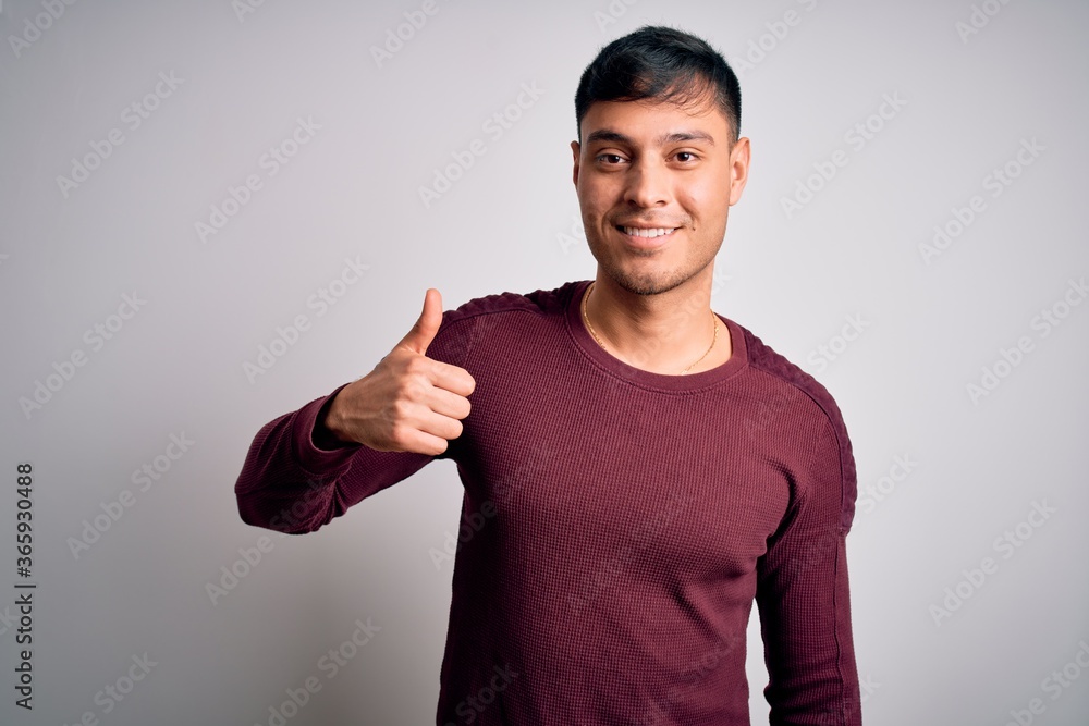 Young handsome hispanic man wearing casual shirt standing over white isolated background doing happy thumbs up gesture with hand. Approving expression looking at the camera showing success. Young handsome hispanic man wearing casual shirt standing over white isolated background doing happy thumbs up gesture with hand. Approving expression looking at the camera showing success.