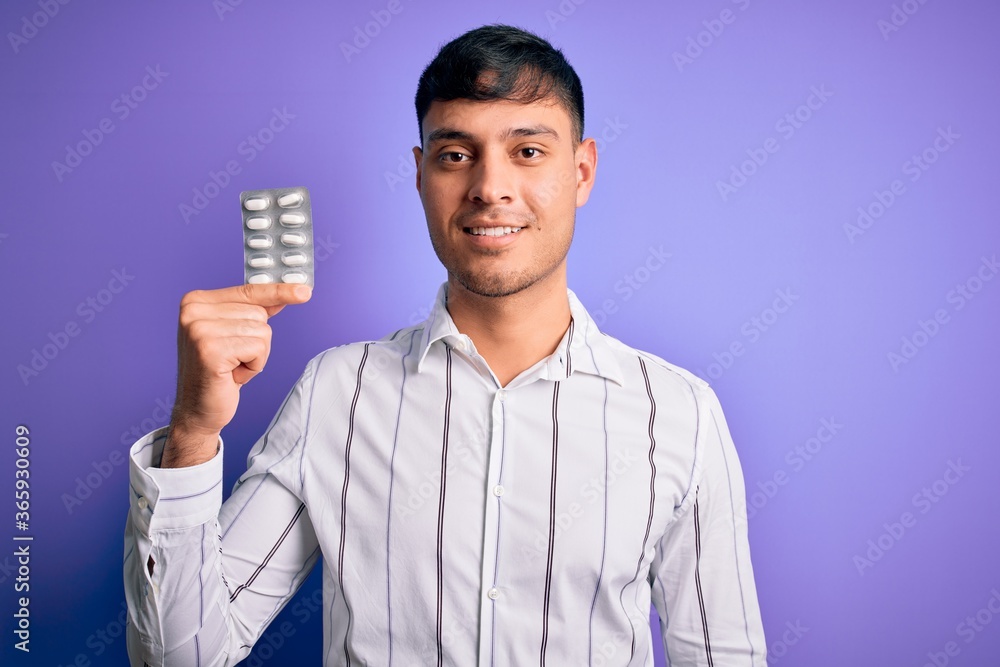 Young hispanic man holding pharmaceutical antibiotics pills over purple isolated background with a happy face standing and smiling with a confident smile showing teeth Young hispanic man holding pharmaceutical antibiotics pills over purple isolated background with a happy face standing and smiling with a confident smile showing teeth