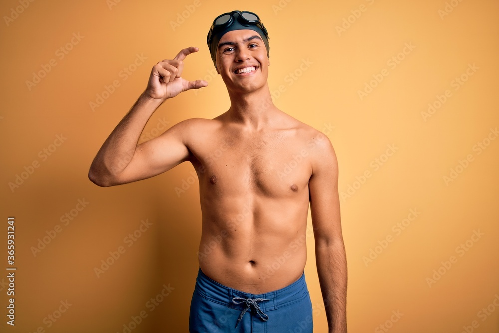 Young handsome man shirtless wearing swimsuit and swim cap over isolated yellow background smiling and confident gesturing with hand doing small size sign with fingers looking and the camera. Measure.