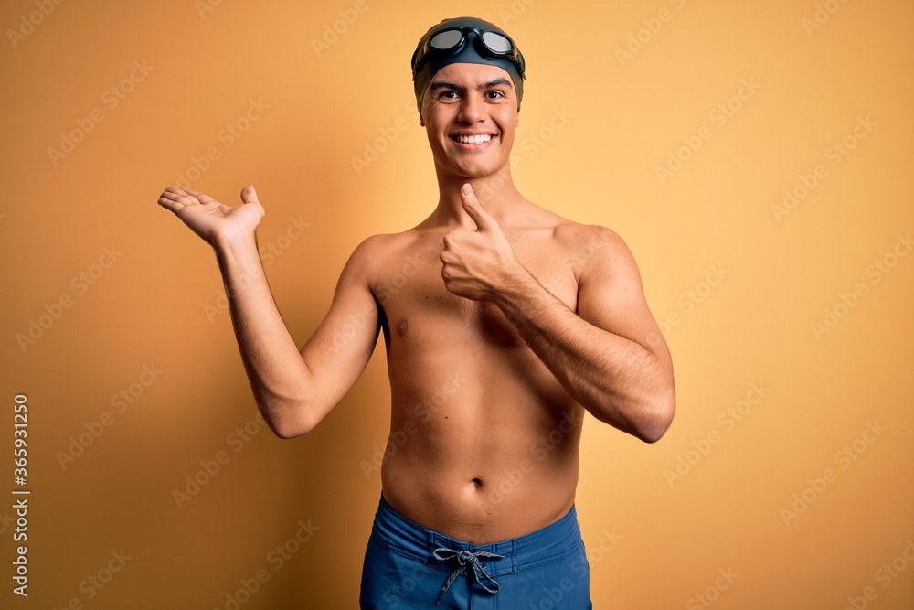 Young handsome man shirtless wearing swimsuit and swim cap over isolated yellow background Showing palm hand and doing ok gesture with thumbs up, smiling happy and cheerful
