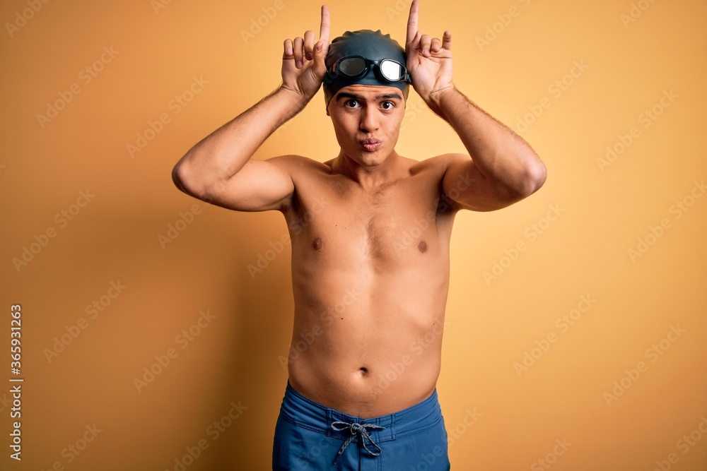 Young handsome man shirtless wearing swimsuit and swim cap over isolated yellow background doing funny gesture with finger over head as bull horns