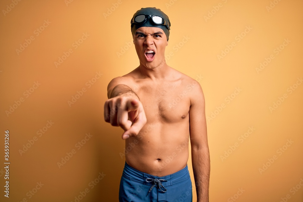 Young handsome man shirtless wearing swimsuit and swim cap over isolated yellow background pointing displeased and frustrated to the camera, angry and furious with you