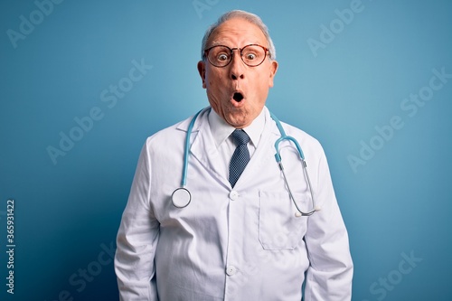 Senior grey haired doctor man wearing stethoscope and medical coat over blue background afraid and shocked with surprise expression, fear and excited face. photo