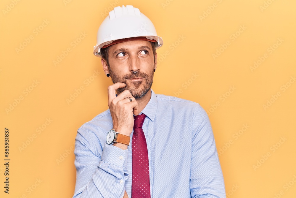 Young handsome man wearing architect hardhat thinking concentrated about doubt with finger on chin and looking up wondering Young handsome man wearing architect hardhat thinking concentrated about doubt with finger on chin and looking up wondering