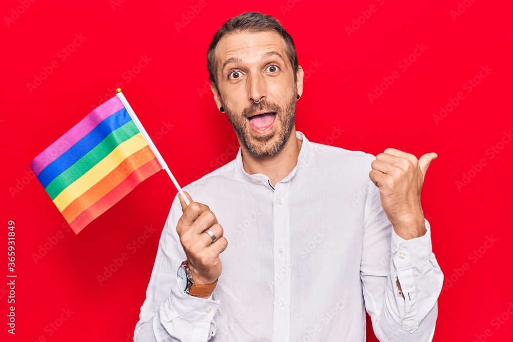 Young handsome man holding rainbow lgbtq flag pointing thumb up to the side smiling happy with open mouth Young handsome man holding rainbow lgbtq flag pointing thumb up to the side smiling happy with open mouth