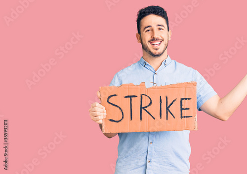 Young hispanic man holding strike banner cardboard celebrating victory with happy smile and winner expression with raised hands