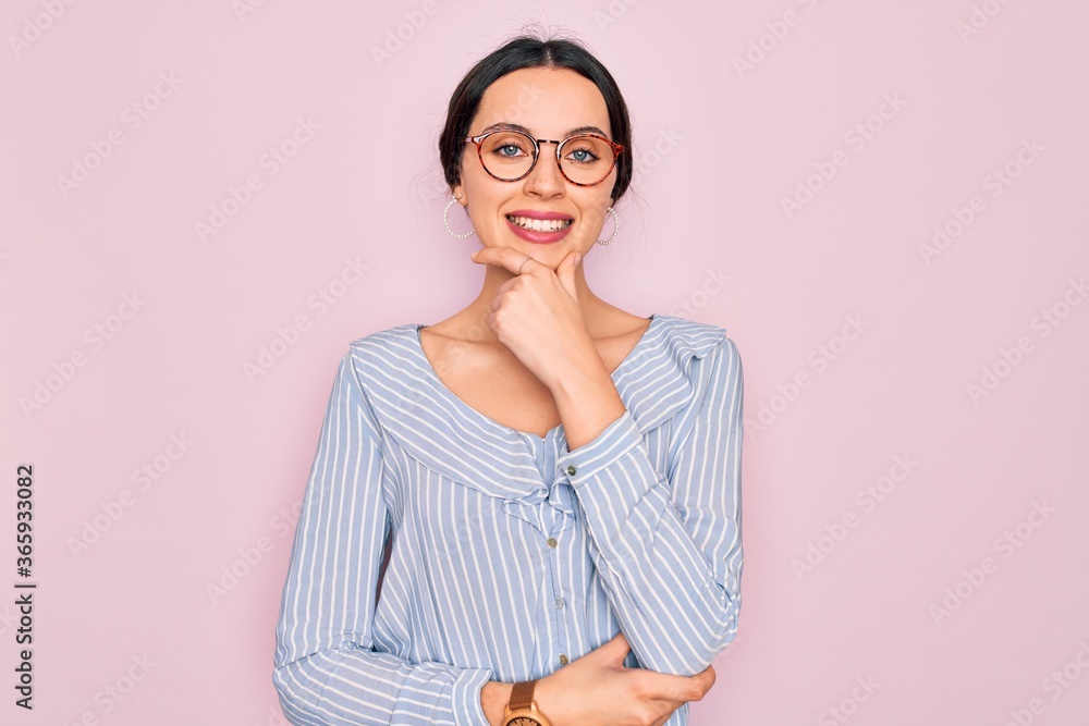 Young beautiful woman wearing casual striped shirt and glasses over pink background looking confident at the camera smiling with crossed arms and hand raised on chin. Thinking positive.