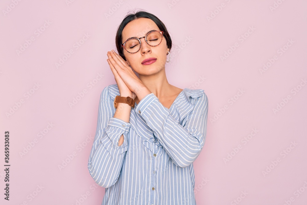 Young beautiful woman wearing casual striped shirt and glasses over pink background sleeping tired dreaming and posing with hands together while smiling with closed eyes.