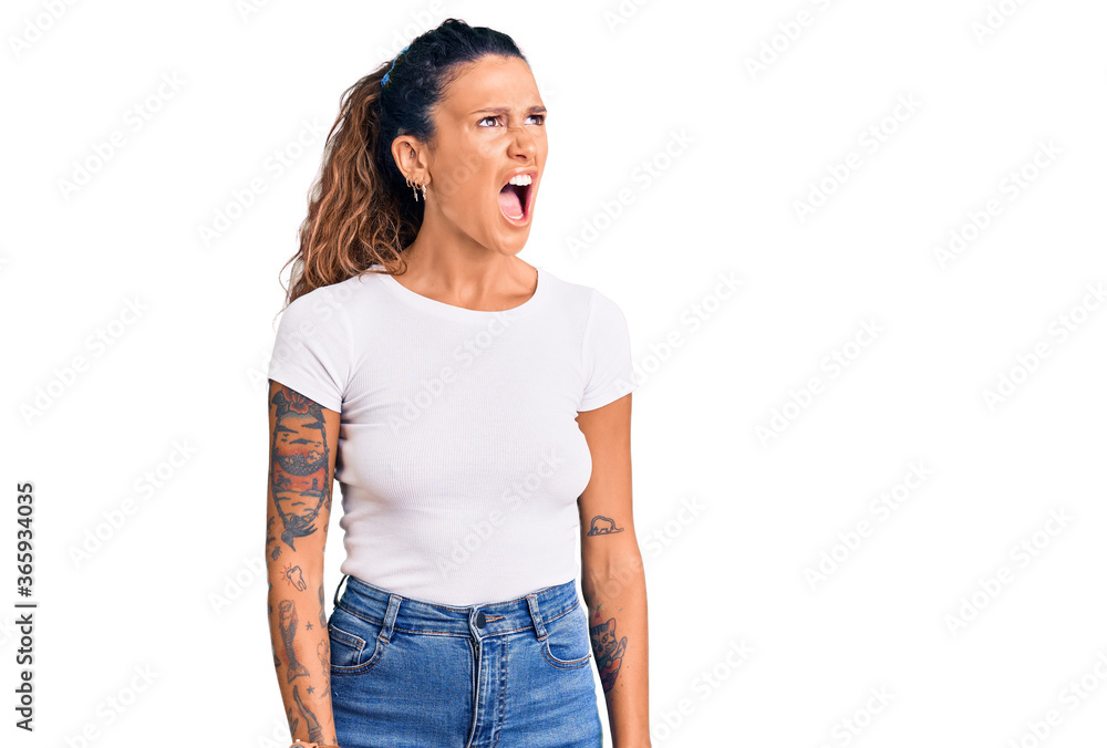 Fototapeta premium Young hispanic woman with tattoo wearing casual white tshirt angry and mad screaming frustrated and furious, shouting with anger. rage and aggressive concept.