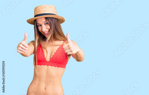 Beautiful brunette young woman wearing bikini approving doing positive gesture with hand, thumbs up smiling and happy for success. winner gesture.