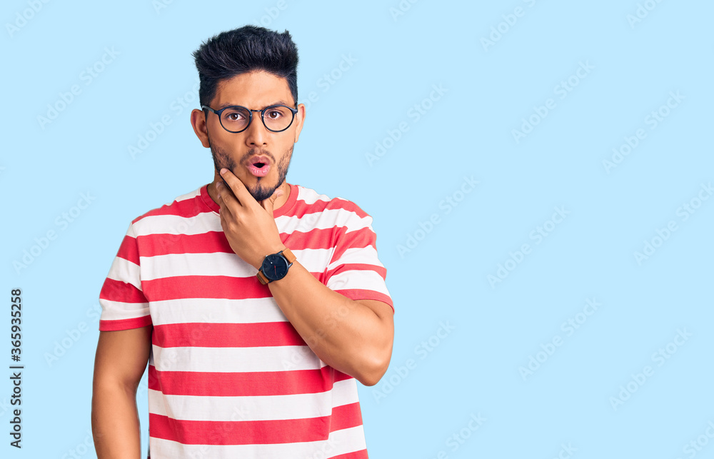 Handsome latin american young man wearing casual clothes and glasses looking fascinated with disbelief, surprise and amazed expression with hands on chin Handsome latin american young man wearing casual clothes and glasses looking fascinated with disbelief, surprise and amazed expression with hands on chin