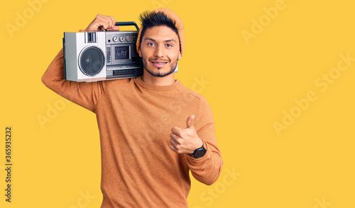 Handsome latin american young man holding boombox, listening to music doing happy thumbs up gesture with hand. approving expression looking at the camera showing success.