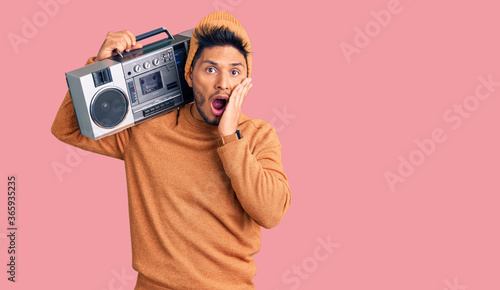 Handsome latin american young man holding boombox, listening to music afraid and shocked, surprise and amazed expression with hands on face