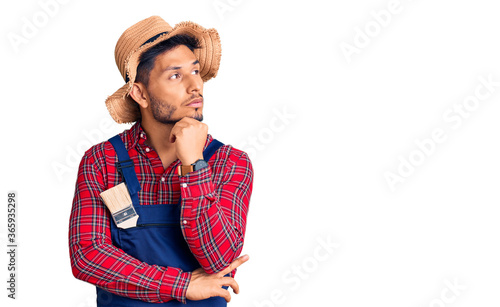 Handsome latin american young man weaing handyman uniform with hand on chin thinking about question, pensive expression. smiling with thoughtful face. doubt concept. photo