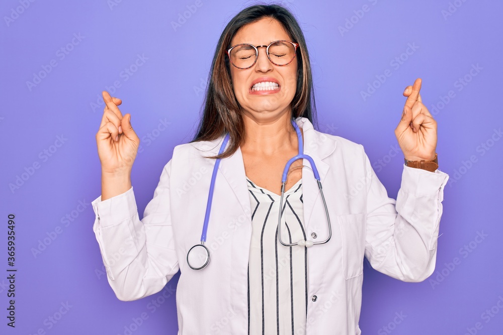 Professional doctor woman wearing stethoscope and medical coat over purple background gesturing finger crossed smiling with hope and eyes closed. Luck and superstitious concept. Professional doctor woman wearing stethoscope and medical coat over purple background gesturing finger crossed smiling with hope and eyes closed. Luck and superstitious concept.