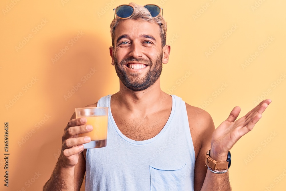Young handsome blond man with beard on vacation drinking glass of healthy orange juice celebrating achievement with happy smile and winner expression with raised hand Young handsome blond man with beard on vacation drinking glass of healthy orange juice celebrating achievement with happy smile and winner expression with raised hand