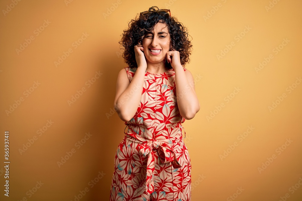 Young beautiful curly arab woman on vacation wearing summer floral dress and sunglasses covering ears with fingers with annoyed expression for the noise of loud music. Deaf concept. Young beautiful curly arab woman on vacation wearing summer floral dress and sunglasses covering ears with fingers with annoyed expression for the noise of loud music. Deaf concept.