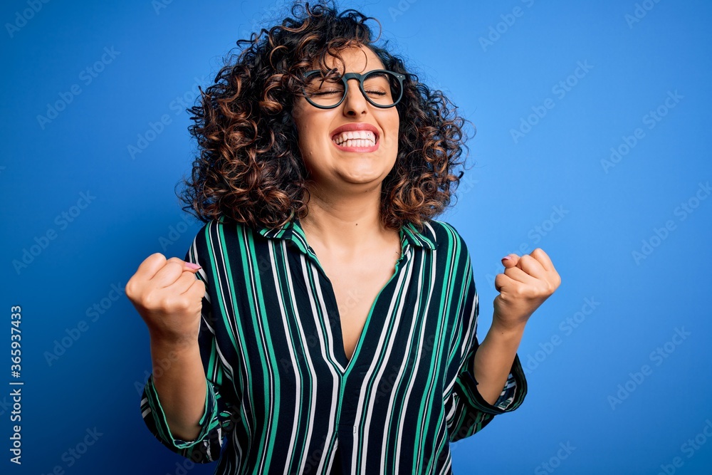 Young beautiful curly arab woman wearing striped shirt and glasses over blue background excited for success with arms raised and eyes closed celebrating victory smiling. Winner concept. Young beautiful curly arab woman wearing striped shirt and glasses over blue background excited for success with arms raised and eyes closed celebrating victory smiling. Winner concept.