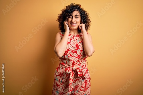 Young beautiful curly arab woman on vacation wearing summer floral dress and sunglasses covering ears with fingers with annoyed expression for the noise of loud music. Deaf concept.
