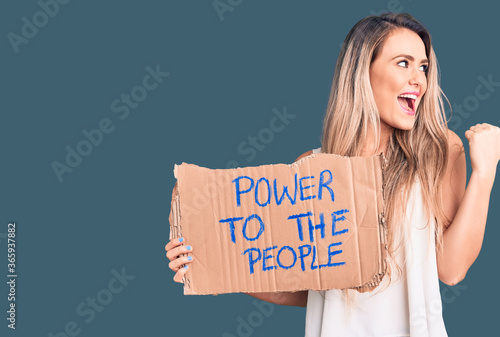 Young beautiful blonde woman holding power to the people cardboard banner pointing thumb up to the side smiling happy with open mouth