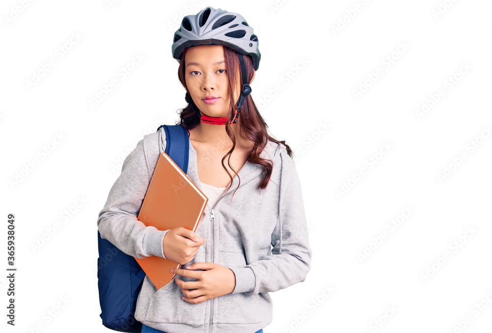 Young beautiful chinese girl wearing student backpack and bike helmet holding book thinking attitude and sober expression looking self confident Young beautiful chinese girl wearing student backpack and bike helmet holding book thinking attitude and sober expression looking self confident