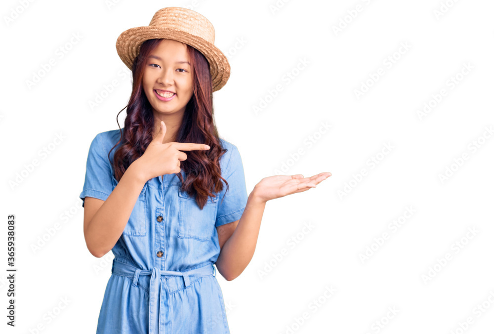 Young beautiful chinese girl wearing summer hat amazed and smiling to the camera while presenting with hand and pointing with finger. Young beautiful chinese girl wearing summer hat amazed and smiling to the camera while presenting with hand and pointing with finger.