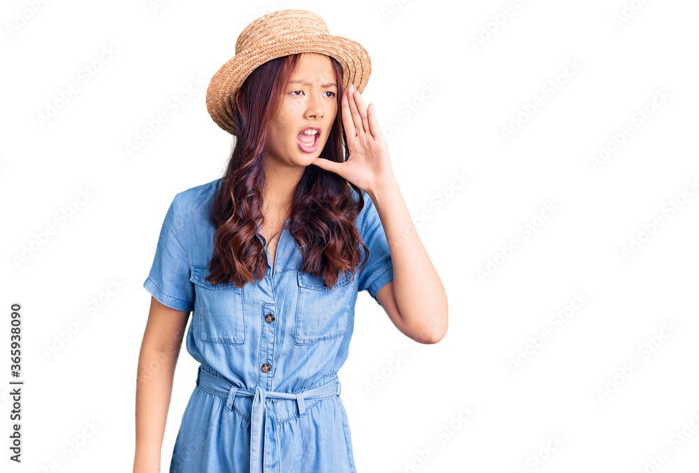 Young beautiful chinese girl wearing summer hat shouting and screaming loud to side with hand on mouth. communication concept. Young beautiful chinese girl wearing summer hat shouting and screaming loud to side with hand on mouth. communication concept.