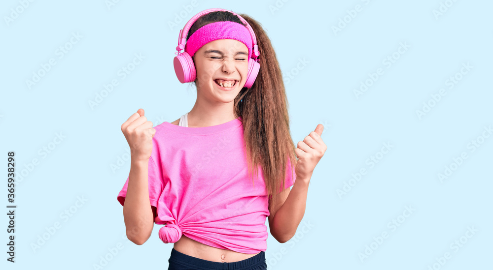Cute hispanic child girl wearing gym clothes and using headphones very happy and excited doing winner gesture with arms raised, smiling and screaming for success. celebration concept. Cute hispanic child girl wearing gym clothes and using headphones very happy and excited doing winner gesture with arms raised, smiling and screaming for success. celebration concept.
