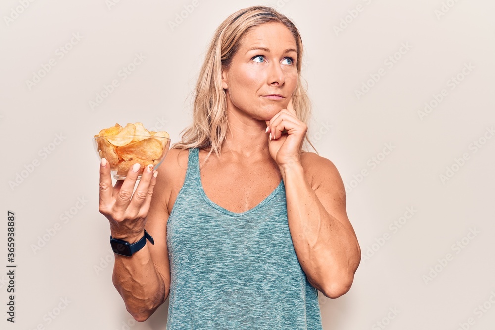 Middle age caucasian blonde woman holding potato chips serious face thinking about question with hand on chin, thoughtful about confusing idea Middle age caucasian blonde woman holding potato chips serious face thinking about question with hand on chin, thoughtful about confusing idea