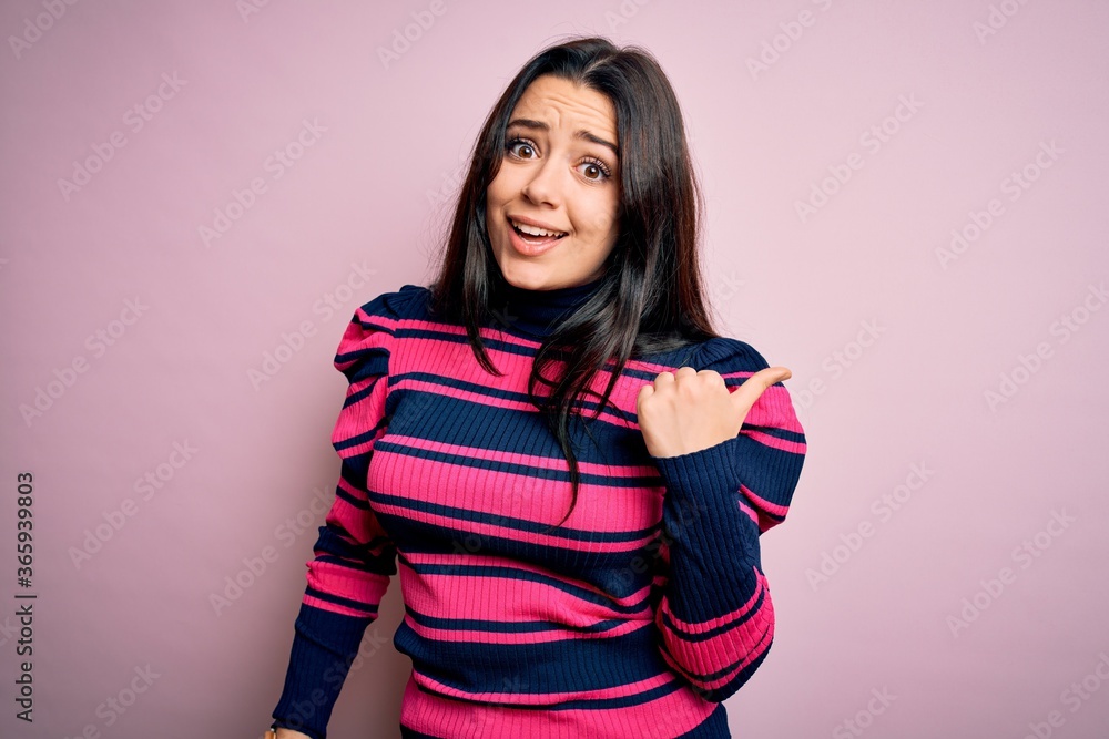 Young brunette elegant woman wearing striped shirt over pink isolated background smiling with happy face looking and pointing to the side with thumb up. Young brunette elegant woman wearing striped shirt over pink isolated background smiling with happy face looking and pointing to the side with thumb up.