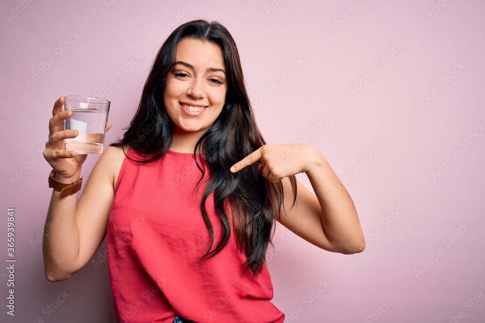Young brunette woman drinking a glass of fresh water over pink isolated background with surprise face pointing finger to himself Young brunette woman drinking a glass of fresh water over pink isolated background with surprise face pointing finger to himself