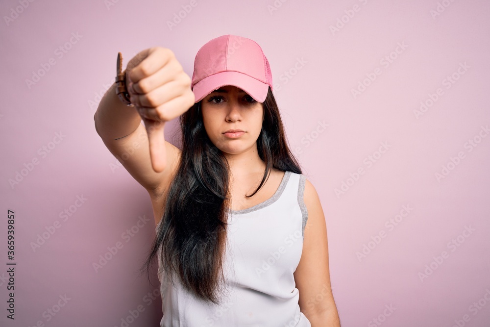 Young brunette woman wearing casual sport cap over pink background looking unhappy and angry showing rejection and negative with thumbs down gesture. Bad expression. Young brunette woman wearing casual sport cap over pink background looking unhappy and angry showing rejection and negative with thumbs down gesture. Bad expression.