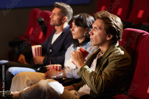 Side view portrait of young people watching movie in cinema while sitting in row on red velvet chairs, focus on man drinking soda through straw with tense face expression, copy space