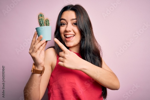 Young brunette woman holding succulent cactus plant over pink background very happy pointing with hand and finger