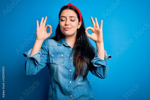 Young brunette woman wearing casual denim shirt over blue isolated background relaxed and smiling with eyes closed doing meditation gesture with fingers. Yoga concept.