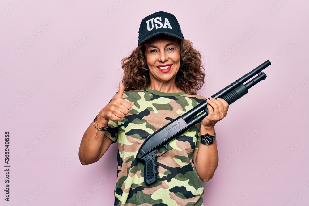 Middle age beautiful hunter woman wearing camouflage t-shirt and usa cap holding shotgun smiling happy and positive, thumb up doing excellent and approval sign Middle age beautiful hunter woman wearing camouflage t-shirt and usa cap holding shotgun smiling happy and positive, thumb up doing excellent and approval sign