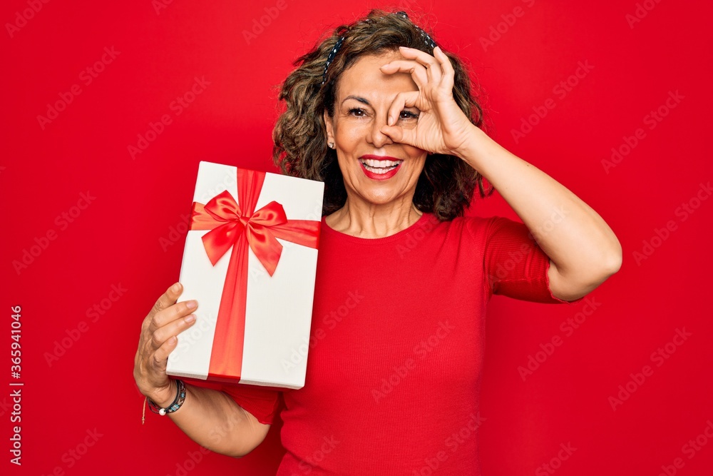 Middle age senior woman holding romantic present box over red isolated background with happy face smiling doing ok sign with hand on eye looking through fingers Middle age senior woman holding romantic present box over red isolated background with happy face smiling doing ok sign with hand on eye looking through fingers