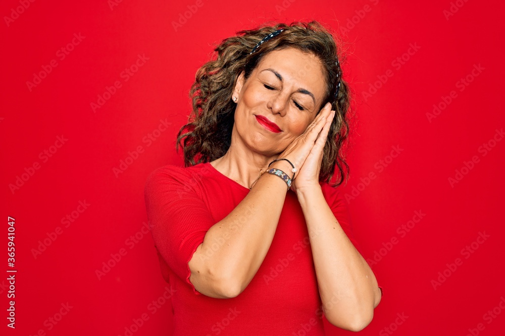 Middle age senior brunette woman wearing casual t-shirt standing over red background sleeping tired dreaming and posing with hands together while smiling with closed eyes.
