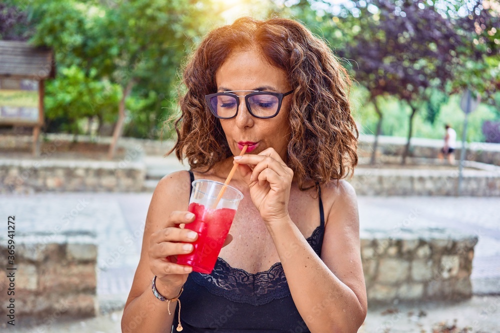 Middle age beautiful woman wearing casual tshirt and glasses. Drinking red cocktail using straw at terrace Middle age beautiful woman wearing casual tshirt and glasses. Drinking red cocktail using straw at terrace