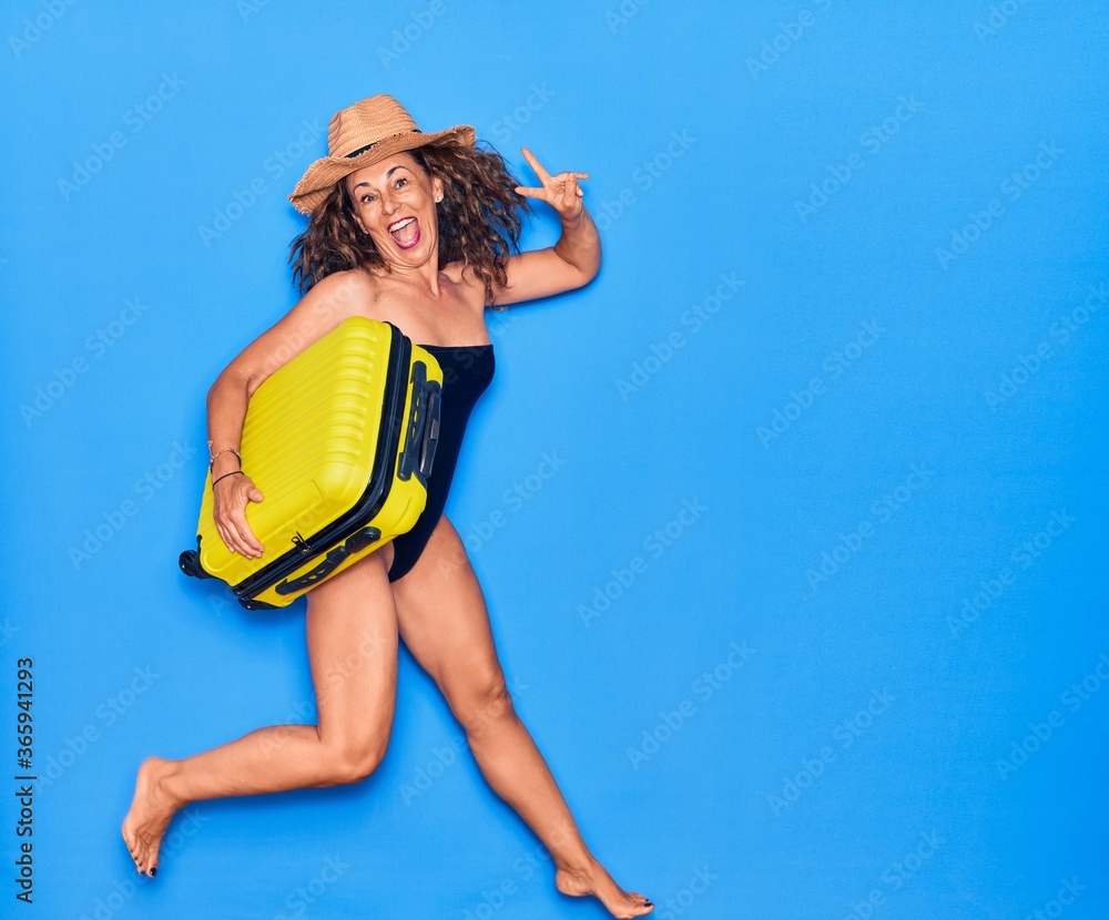 Middle age beautiful woman on vacation wearing swimwear and hat smiling happy. Jumping with smile on face holding cabin bag over isolated background Middle age beautiful woman on vacation wearing swimwear and hat smiling happy. Jumping with smile on face holding cabin bag over isolated background