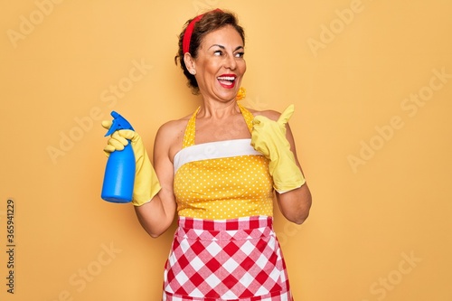 Middle age senior housewife pin up woman wearing 50s style retro dress cleaning using spray pointing and showing with thumb up to the side with happy face smiling
