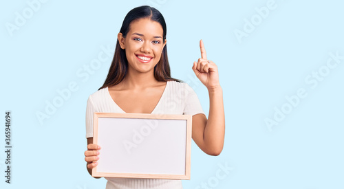 Young beautiful latin girl holding empty white chalkboard surprised with an idea or question pointing finger with happy face, number one