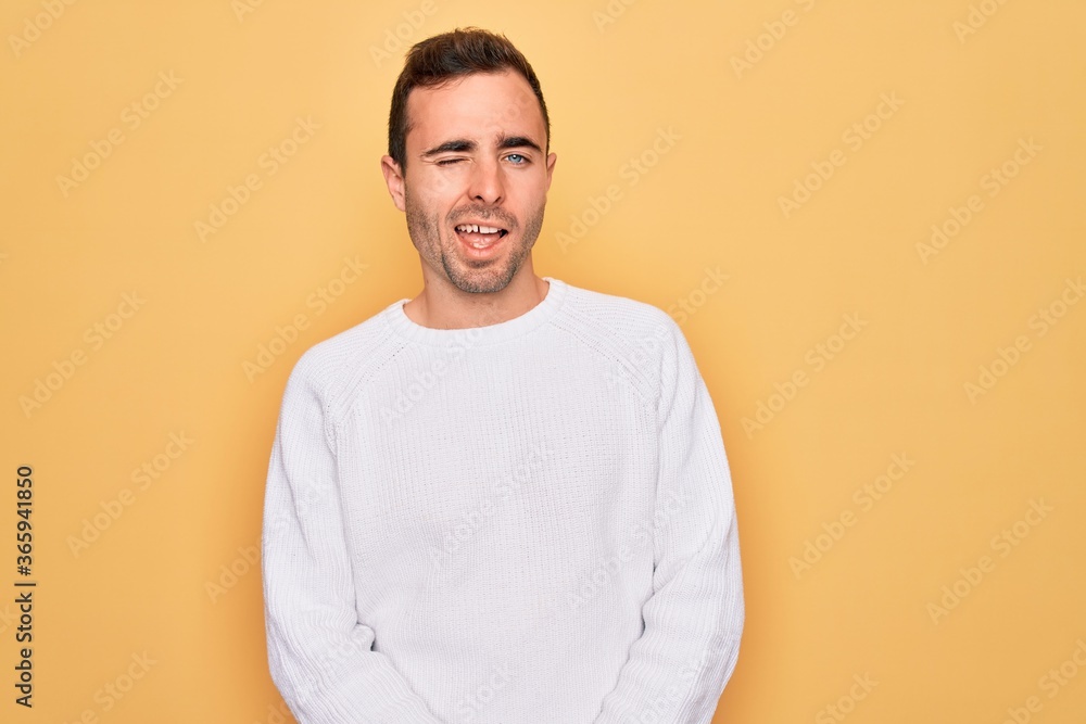 Young handsome man with blue eyes wearing casual sweater standing over yellow background winking looking at the camera with sexy expression, cheerful and happy face. Young handsome man with blue eyes wearing casual sweater standing over yellow background winking looking at the camera with sexy expression, cheerful and happy face.