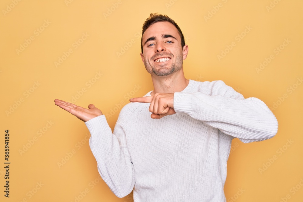 Young handsome man with blue eyes wearing casual sweater standing over yellow background amazed and smiling to the camera while presenting with hand and pointing with finger. Young handsome man with blue eyes wearing casual sweater standing over yellow background amazed and smiling to the camera while presenting with hand and pointing with finger.
