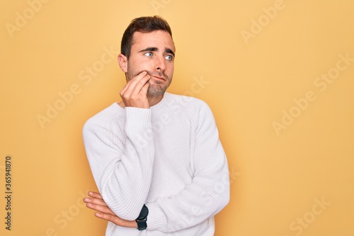 Young handsome man with blue eyes wearing casual sweater standing over yellow background with hand on chin thinking about question, pensive expression. Smiling with thoughtful face. Doubt concept. © Krakenimages.com