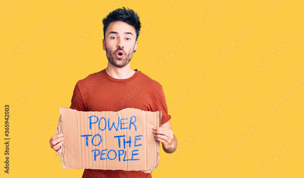 Young hispanic man holding power to the people banner scared and amazed with open mouth for surprise, disbelief face Young hispanic man holding power to the people banner scared and amazed with open mouth for surprise, disbelief face